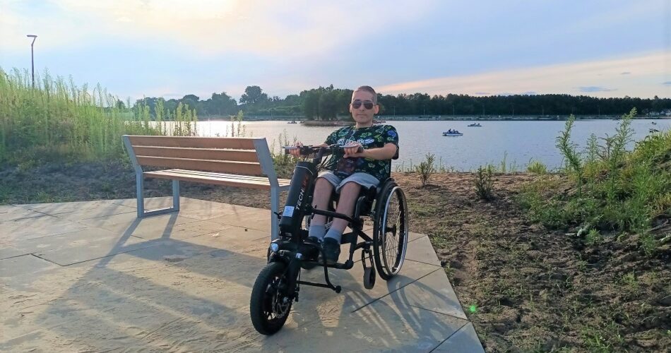 Autor bloga - Paweł Borowiecki korzystający z wózka z przystawką elektryczną. W tle zachód słońca nad jeziorem.