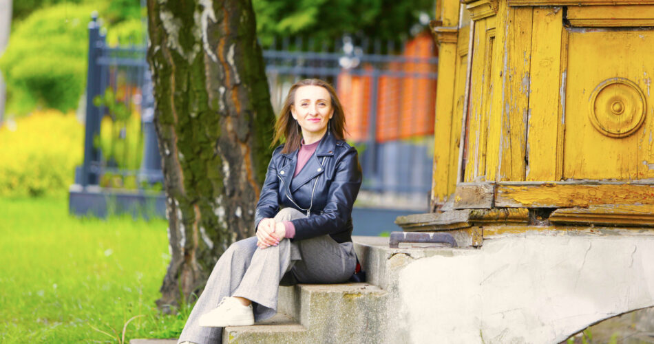 Zdjęcie przedstawia Anetę Kalbarczyk siedzącą na schodkach przed domem. W tle drzewo.
