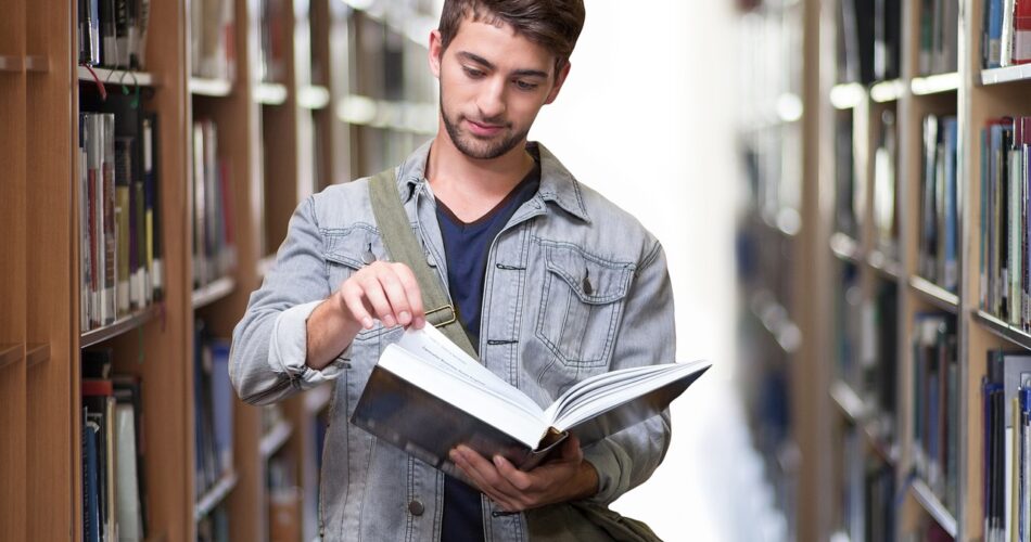 Zdjęcie przedstawia nauczyciela w bibliotece. Trzyma w rękach książkę. W tle znajdują się regały z książkami.